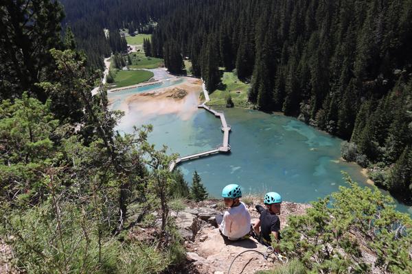 Two individuals wearing helmets sit on a rocky mountain ledge, gazing down at a stunning turquoise lake nestled within a dense coniferous forest